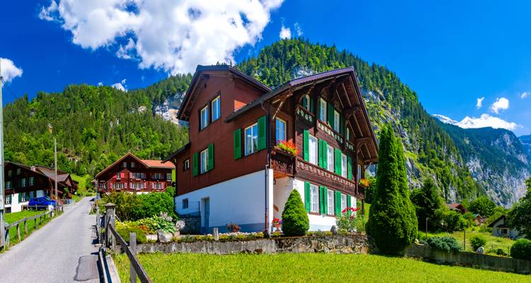 Traditionelles Holz-Alpenhaus mit grünen Fensterläden, eingebettet in saftige Wiesen und umgeben von ragenden Bergen unter einem strahlend blauen Himmel.