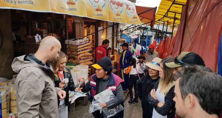 Les voyageurs interagissent avec un vendeur local dans un marché en plein air animé, entourés de collations et de bâches colorées.