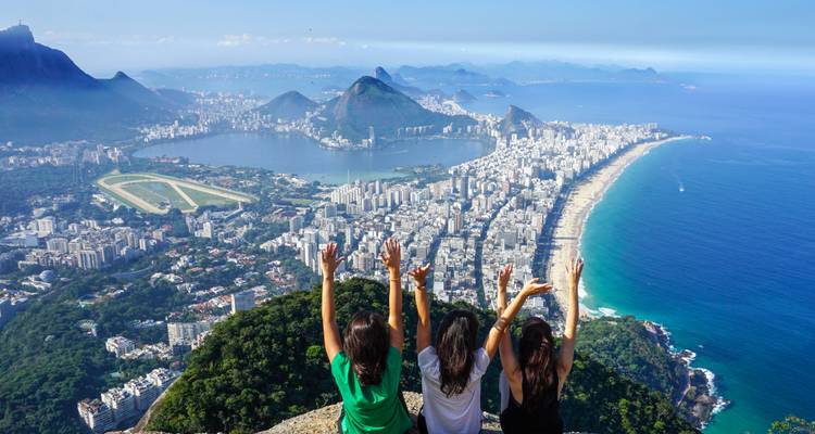 Tres mujeres jóvenes levantando los brazos en celebración en la cima con vista a Río de Janeiro, laguna y playas.
