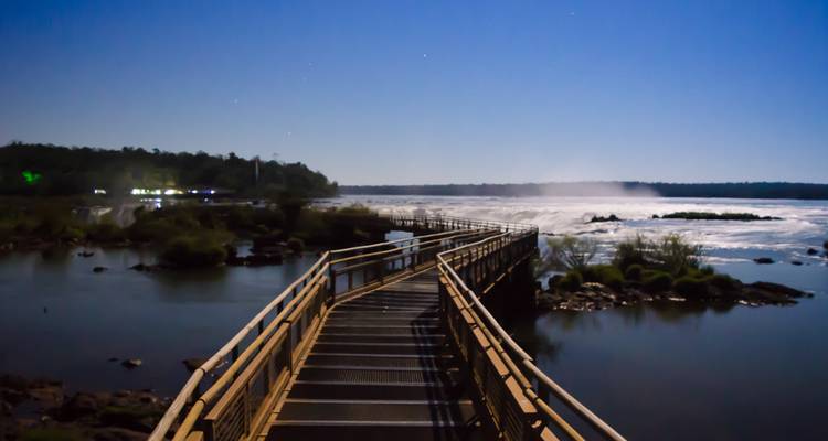 Fotografía de larga exposición nocturna de un pasarela elevada que conduce hacia las brumosas Cataratas del Iguazú bajo un cielo estrellado.