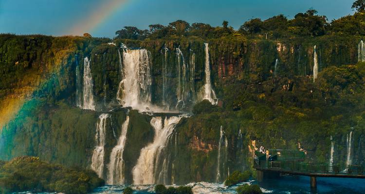 Las espectaculares Cataratas del Iguazú enmarcadas por un arcoíris vibrante y una exuberante vegetación con visitantes en una plataforma de observación.