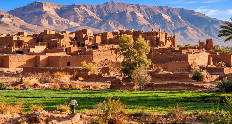 Las murallas de adobe del pueblo se mezclan con el árido paisaje montañoso, mientras un solitario granjero cultiva un campo verde en primer plano.