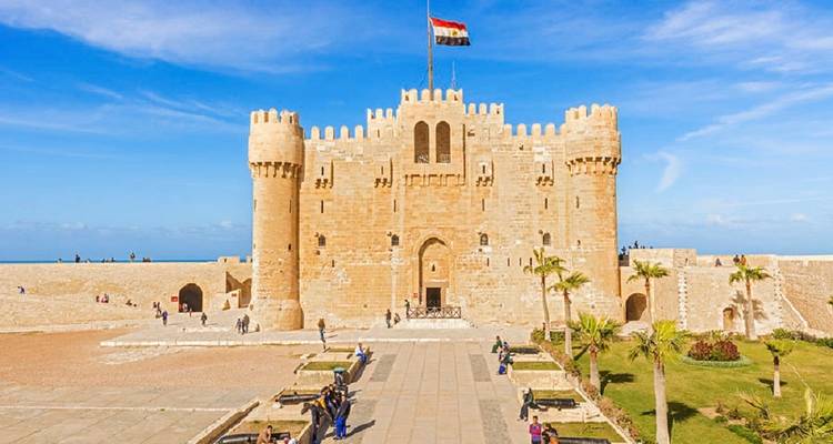 Vue de face de la Citadelle de Qaitbay avec le drapeau égyptien flottant au-dessus de deux tours jumelles par une journée ensoleillée.