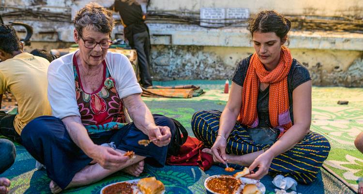 Deux femmes sont assises sur un tapis au sol aux motifs variés et partagent un repas de rue épicé servi sur des assiettes en papier.