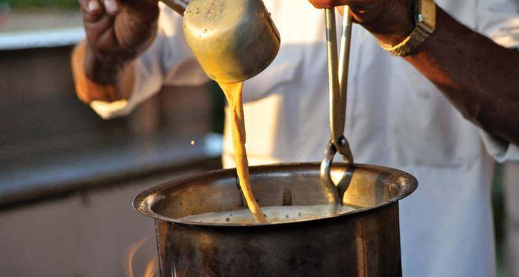 Un chef verse avec dextérité du thé chai fumant d'une louche dans un pot en métal.