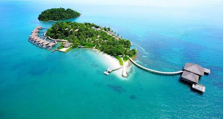 Luftaufnahme einer üppigen Privatinsel, umgeben von klarem türkisfarbenem Meer mit Überwasser-Bungalows und einem geschwungenen Steg.