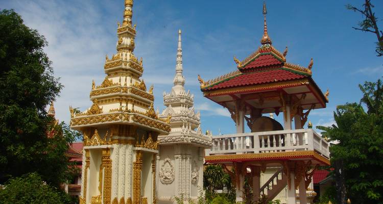 Weiße und goldene Stupas sowie ein rotgedecktes Glockenpavillon inmitten üppigen Grüns unter einem klaren blauen Himmel.