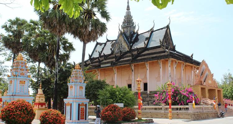 Traditioneller Tempel mit kunstvollen dunklen Dachziegeln und pastellfarbenen Säulen, umrahmt von blühenden Bäumen; eine Person auf einem Motorrad fährt davor vorbei.