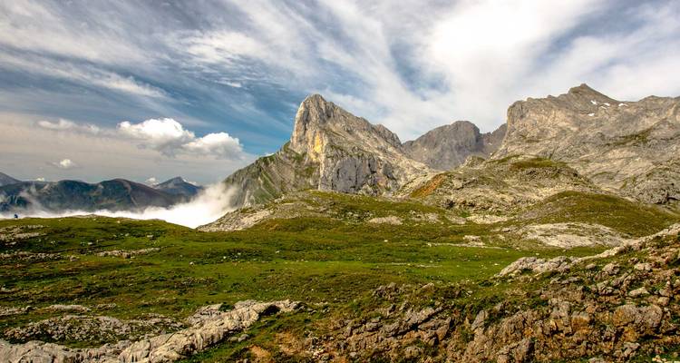 Weitläufige Alpenlandschaft mit zerklüfteten Gipfeln und sanften grünen Wiesen unter einem dynamischen Himmel.