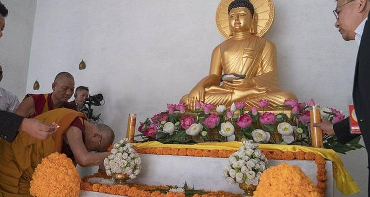 Des moines et des visiteurs rendant hommage devant un autel couronné d'une statue de Bouddha dorée ornée de fleurs roses et blanches.
