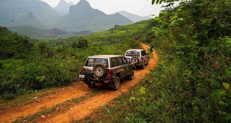Deux véhicules tout-terrain circulent sur une piste de terre rougeâtre à travers des collines verdoyantes luxuriantes et des pics de calcaire lointains dans le nord du Vietnam.
