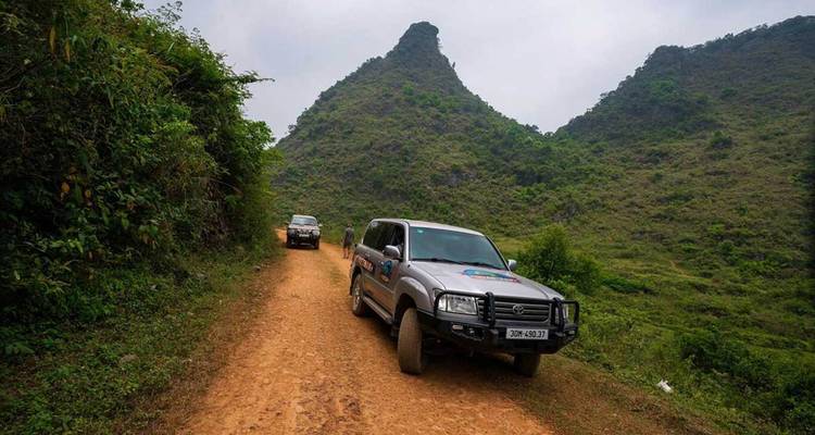 Un camion 4x4 circule sur une route de terre étroite bordée de collines karstiques vertes aux pentes abruptes.