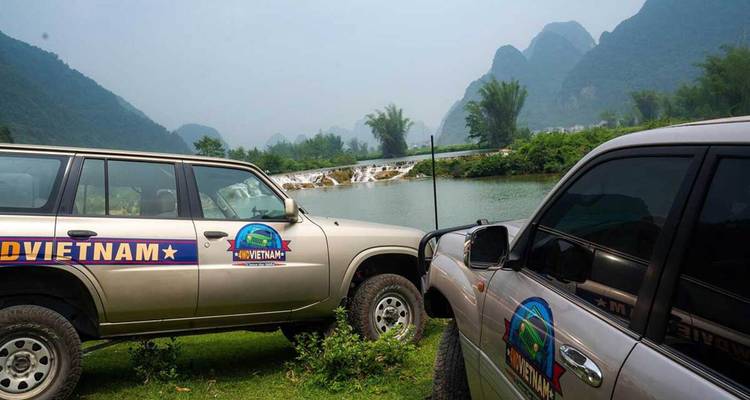 Deux véhicules utilitaires sport sont garés au bord d'une rivière tranquille et d'une petite cascade au milieu de montagnes calcaires enveloppées de brume.