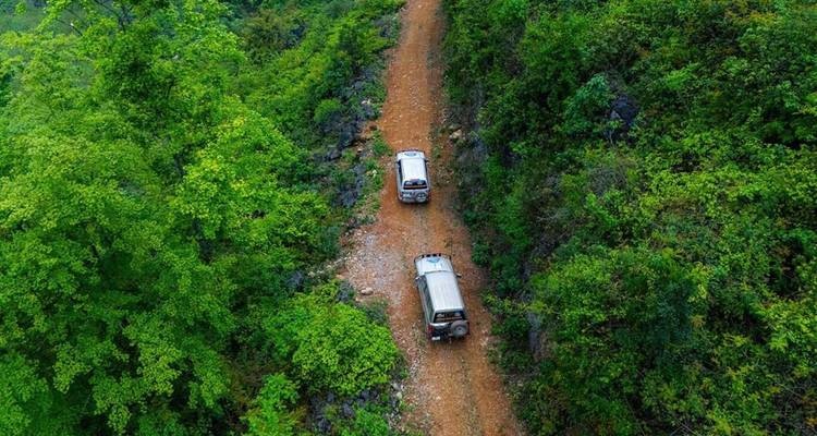 Vue aérienne d'une route en terre orange étroite coupant à travers une jungle verte dense avec deux jeeps en convoi.