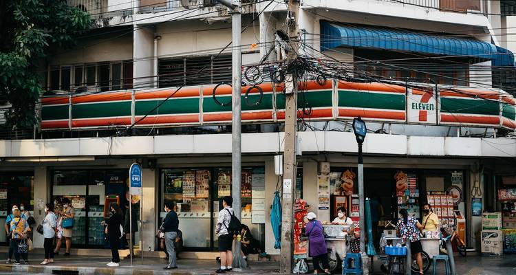 Hoekje in Bangkok met een 7-Eleven winkel, verwarde stroomkabels en gemaskerde voetgangers die in de rij staan bij verkopers.
