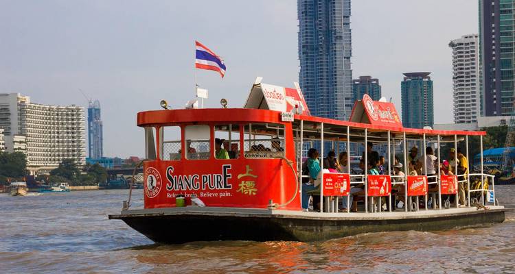 Een rood-wit forenzenbootje vol passagiers vaart over de Chao Phraya-rivier met Bangkoks skyline op de achtergrond.
