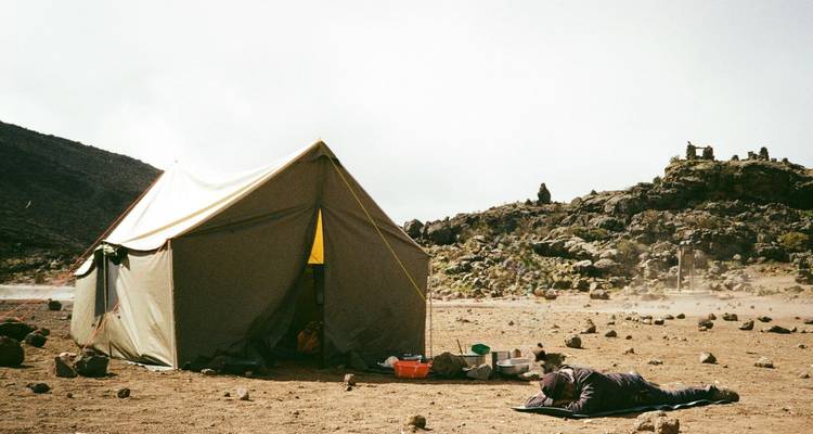 A lone canvas tent stands on a barren alpine plateau while a trekker rests on a mat nearby.