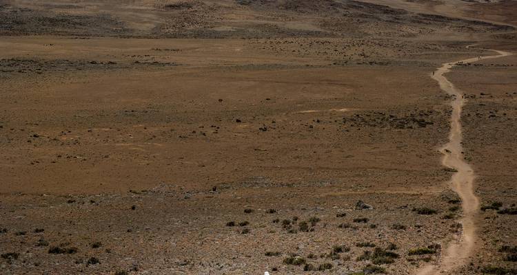 A narrow dusty trail stretches across an expansive barren plateau under muted light.