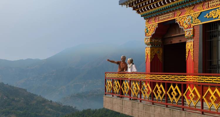 Pareja de adultos mayores de pie en el colorido balcón de un templo bhutanés con vistas a montañas brumosas.