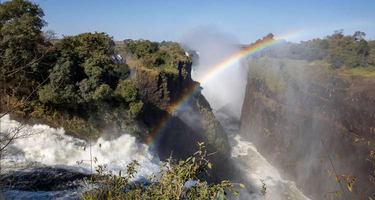 Une puissante cascade se jetant dans un profond canyon avec un arc-en-ciel éclatant arqué au-dessus des embruns, entouré de falaises verdoyantes et luxuriantes.
