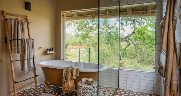Salle de bain élégante de lodge de safari dotée d'une baignoire autoportante, de serviettes jumelées et d'une grande fenêtre donnant sur une brousse verdoyante.