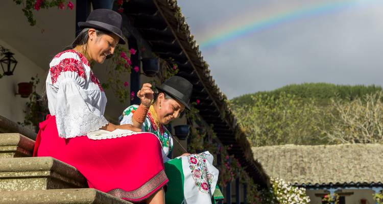 Zwei Frauen in leuchtend traditioneller Kleidung spinnen Garn auf Steinstufen unter einem Regenbogen und blühenden Blumen.
