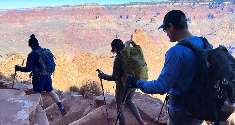 Three backpackers descend a steep switchback with vast canyon vistas stretching beyond.