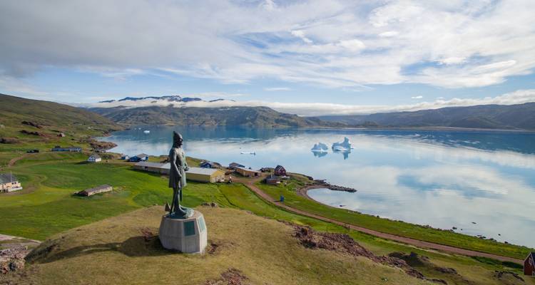 Statue of an explorer overlooks a serene Greenlandic fjord dotted with icebergs and coastal hamlet.