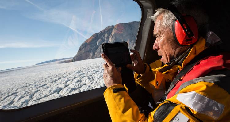 Elderly man in a yellow expedition jacket photographs a vast icy glacier from a helicopter window.