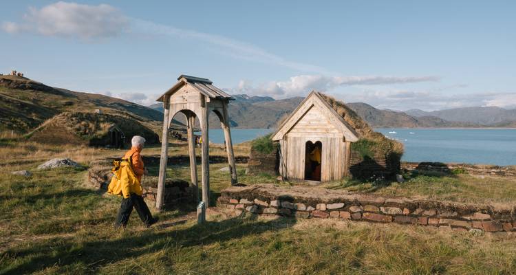 Traveler in yellow jacket explores turf-roofed Norse ruins beside a calm fjord.