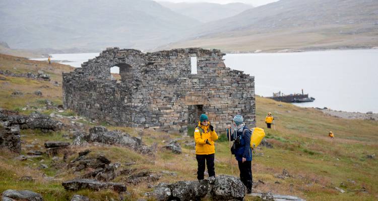 Two hikers pause near ancient stone ruins on a grassy slope above a fjord.