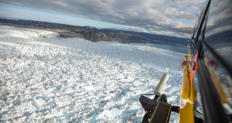 Helicopter flies above a vast rumpled glacier stretching toward distant mountains and sea.
