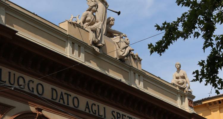 Ligne de toit du théâtre néoclassique ornée de figures sculptées et d'un ange tenant une trompette sur un ciel bleu.