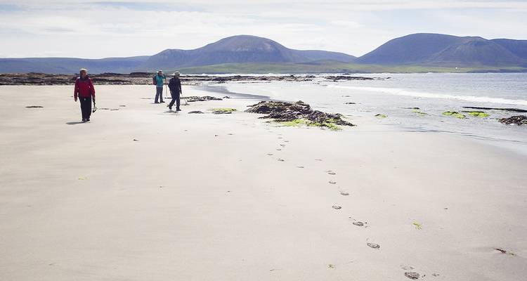 Les promeneurs laissent des empreintes de pas sur une large plage déserte, encadrée par des collines lointaines.