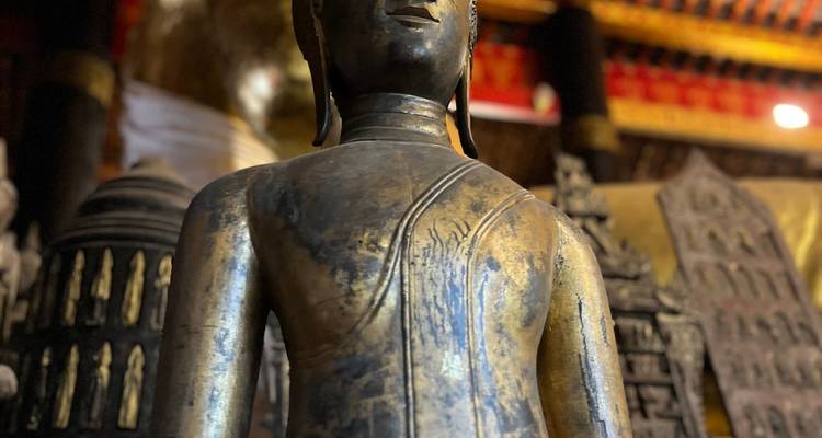Weathered bronze Buddha statue inside a dimly lit temple hall with red and gold décor.