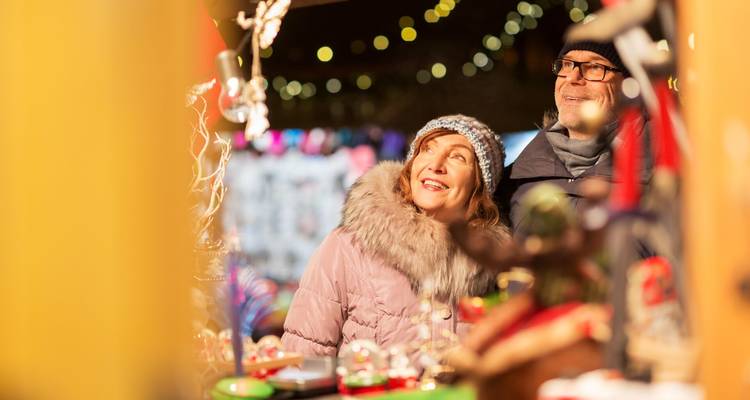 Couple âgé souriants vêtus de vêtements d'hiver admirant un étal de marché de Noël brillamment illuminé avec des lumières festives en arrière-plan.