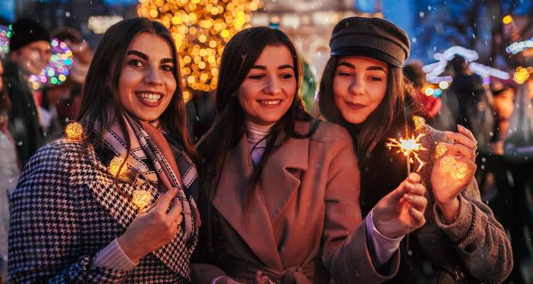 Trois jeunes femmes tenant des cierges magiques et des ornements décorés de guirlandes lumineuses, célébrant dans un marché en plein air festif.