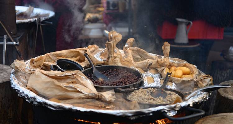 Grand plat rond contenant un assortiment de haricots, de céréales et de boulettes de pâte en cuisson sur une flamme vive à un étal en plein air.