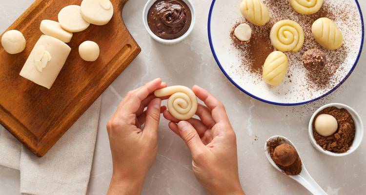 Gros plan sur les mains roulant des spirales de pâte d'amande sur un plan de travail de cuisine avec des bols de chocolat et de cacao à proximité.