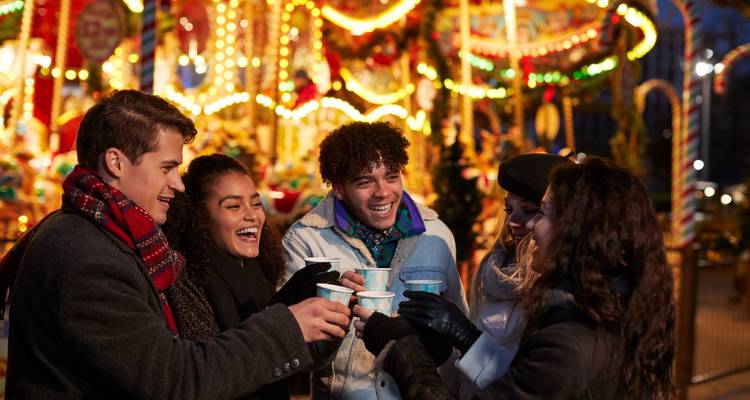 Groupe de jeunes adultes enveloppés dans des écharpes et des manteaux, acclamant avec des boissons chaudes devant un carrousel illuminé.