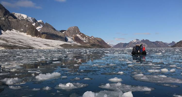 Small inflatable boat navigating through floating sea ice with jagged Arctic mountains in the distance.