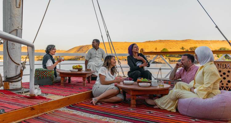 Groupe de voyageurs se détendant sur des tapis et des tables basses à bord d'un voilier traditionnel du Nil au coucher du soleil.