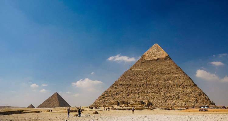 Les trois pyramides de Giza s'élèvent du désert avec des touristes et des chameaux au premier plan sous un ciel bleu profond.