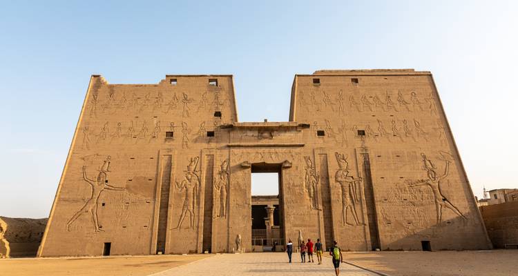 Façade monumentale en grès du Temple d'Edfou avec des reliefs sculptés, les minuscules visiteurs éclipsés à l'entrée.