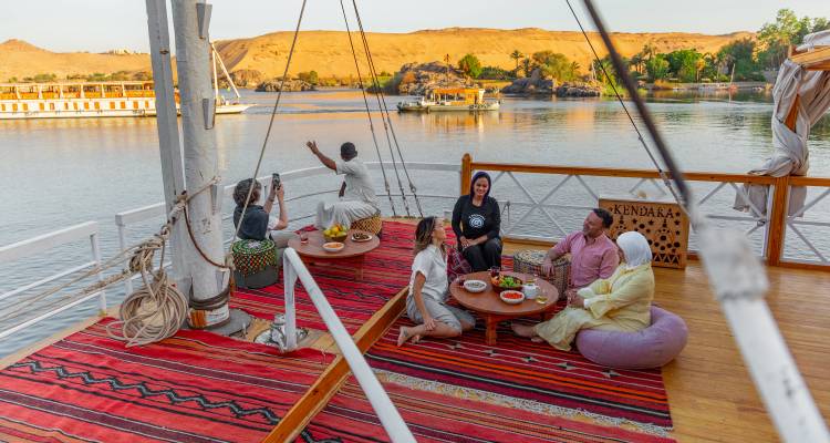 Les voyageurs se détendent sur le pont d'un bateau fluvial du Nil en savourant des boissons avec des paysages désertiques en arrière-plan.