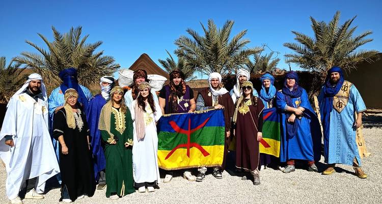 Groupe de touristes vêtus de robes berbères traditionnelles aux couleurs éclatantes posant avec le drapeau amazighe parmi les palmiers.