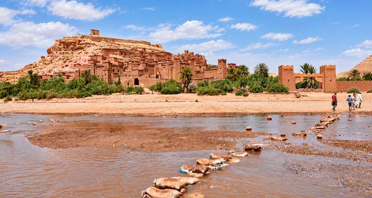 Complejo de ksar de tierra junto a un río poco profundo con turistas cruzando sobre piedras de paso bajo un cielo parcialmente nublado.