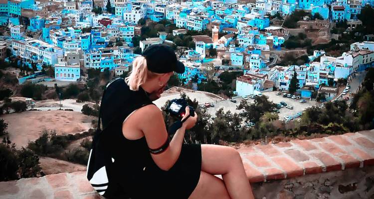 Reiziger zit op een rand en fotografeert het uitgestrekte, blauw geschilderde stadje dat zich onder hem uitstrekt.