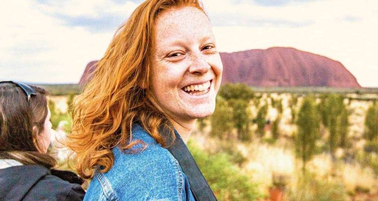 Femme souriante en veste en denim avec le monolithe rouge d'Uluru en arrière-plan.