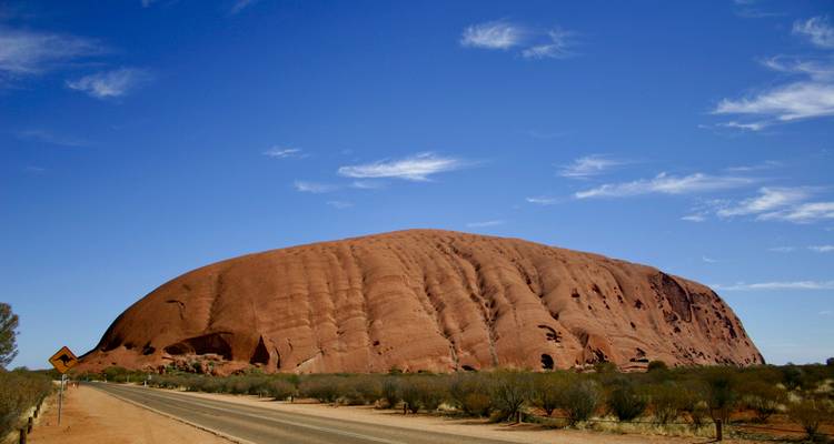 Une longue route rectiligne de l'outback s'approche du flanc étendu d'Uluru sous un ciel strié de nuages.